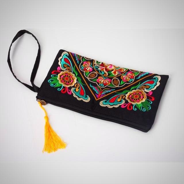The image is of the Galsang Flower Embroidered Clutch. Features: The material is for interior and exterior is cotton, the style is a day clutch, the closure is a zipper, the size is approximately 15cm x 27cm, no straps, interior compartment, embroidered pattern, yellow tassel on the end of the zipper.